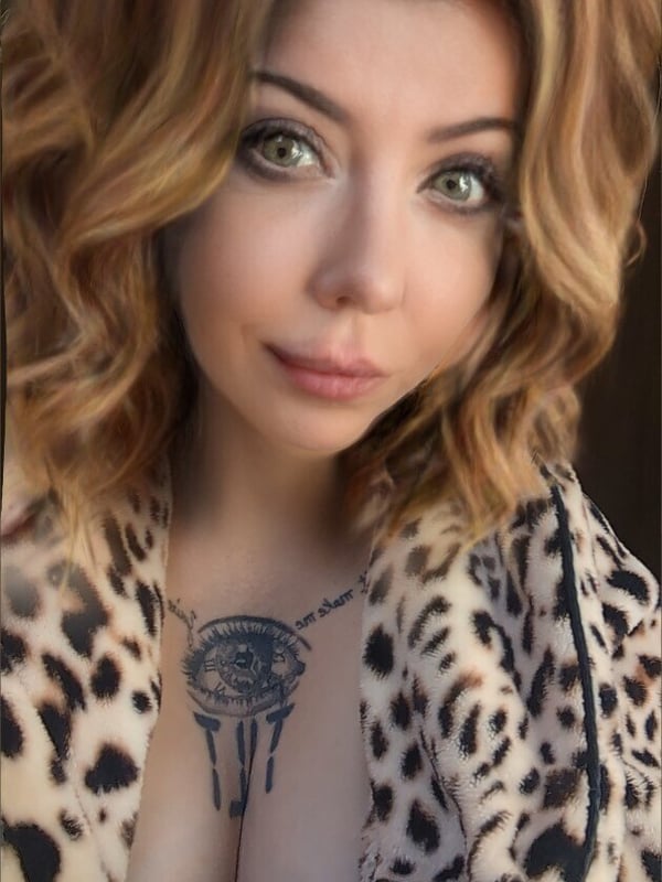 Close-up portrait of a woman with wavy brunette hair, green eyes, and tattoos visible on her chest, wearing a leopard print top.