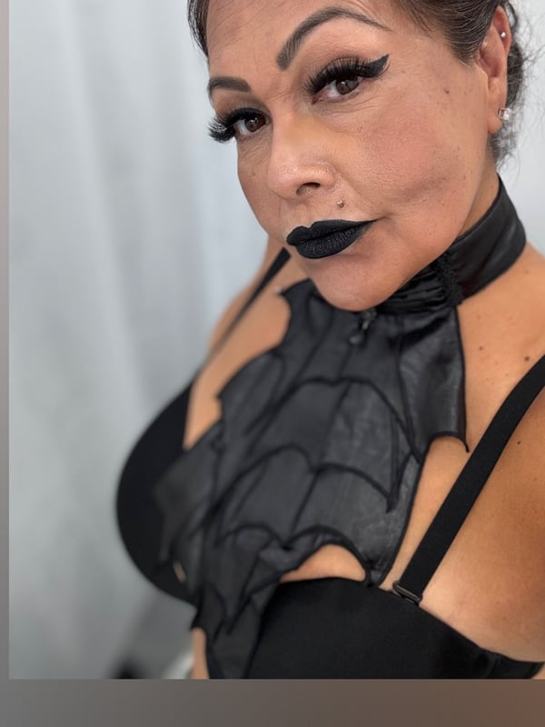 Close-up portrait of a woman with dramatic makeup, wearing a black leather bat-wing harness and choker.