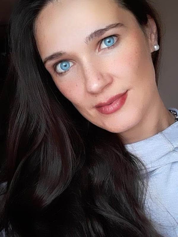 Close-up portrait of a woman with long dark hair and blue eyes, smiling softly while wearing a light top.