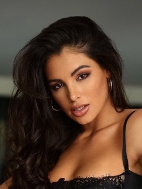 Close-up portrait of a woman with long wavy dark hair, hoop earrings, and bold makeup, wearing a black lace top.