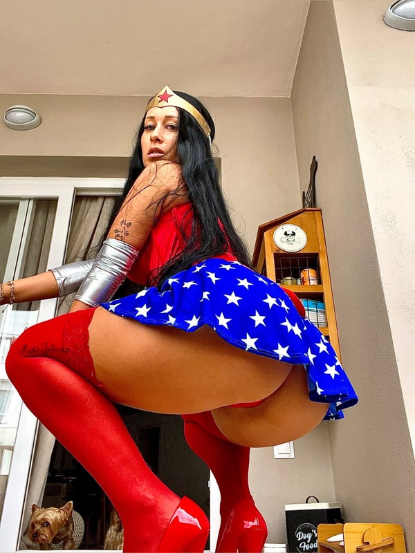 A woman dressed as Wonder Woman in a costume with a star-spangled skirt and red boots, posing dynamically on a balcony with home decor in the background.
