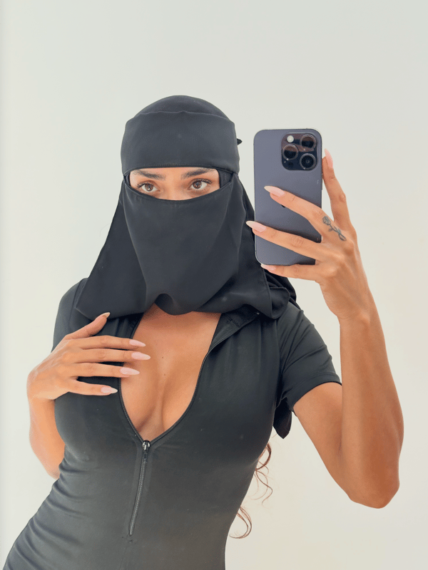 A woman in a white headscarf and blue top stands indoors with a neutral background.