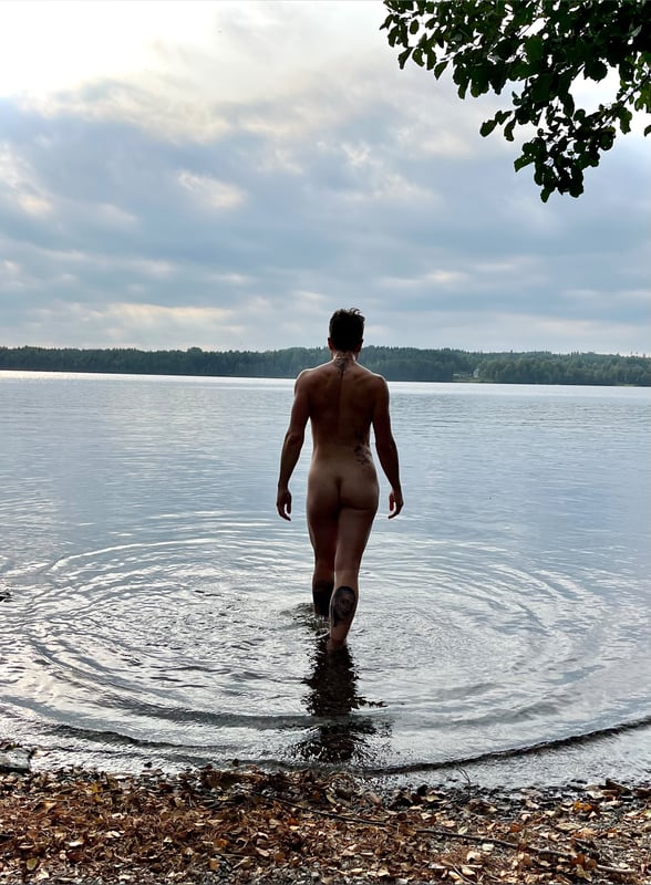 A person standing shirtless in shallow water at a lakeside with trees and a cloudy sky in the background.