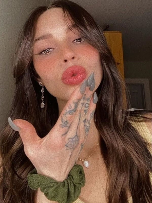 A woman with long brown hair making a kiss face and peace sign, displaying hand tattoos, in a casual indoor setting.