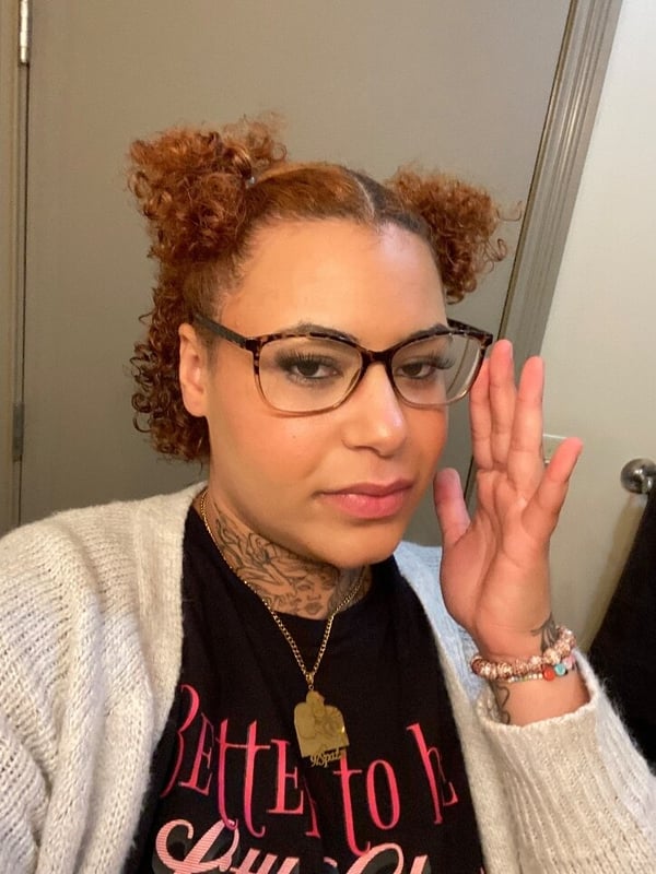 A woman with curly reddish hair in pigtails, wearing glasses and a cardigan over a black top, making a hand gesture indoors.
