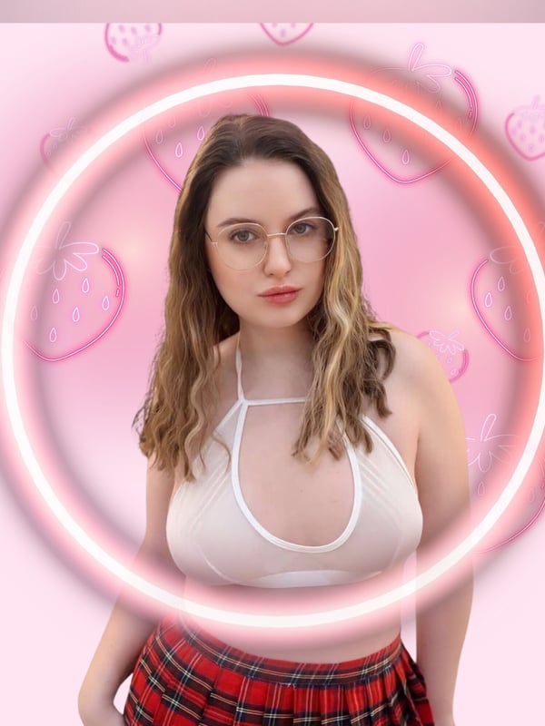 A woman with glasses and wavy brown hair, smiling in a sheer top and plaid skirt against a pink background with heart effects.