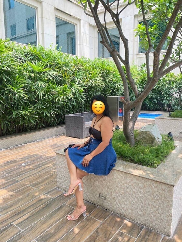 A woman sitting on a tiled poolside area in a blue strapless top and short skirt, with high-heeled sandals, smiling with an emoji over her face, against a backdrop of lush greenery and architecture.