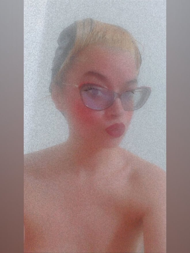 Close-up portrait of a young woman with blonde hair, purple glasses, and red lipstick, smiling softly against a foggy background.