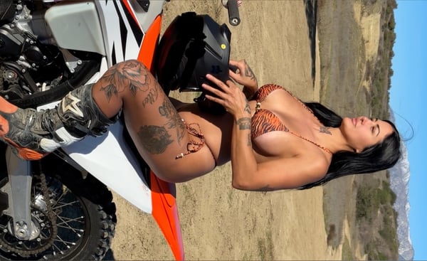 A woman in a bikini posing on a dirt bike in a desert landscape with mountains in the background.