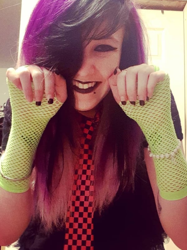 A young woman with vibrant purple and black hair smiling playfully while wearing green mesh arm warmers, a checkered tie, and dark clothing in a casual indoor room.