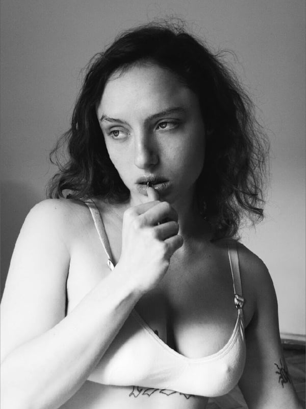 A black and white portrait of a woman with dark wavy hair wearing a light top, posing with her hand near her mouth.