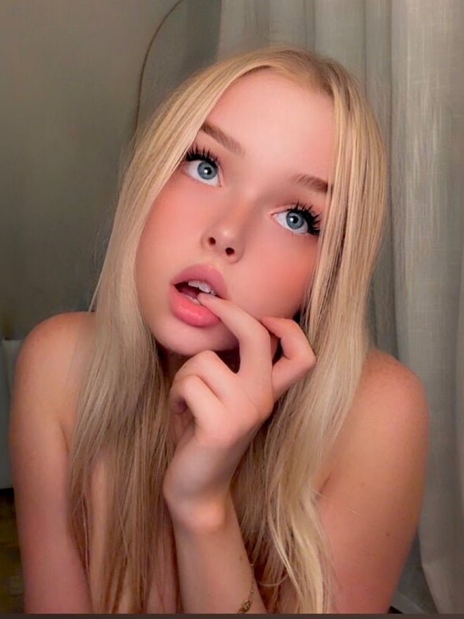 A blonde woman posing indoors with a hand near her face.