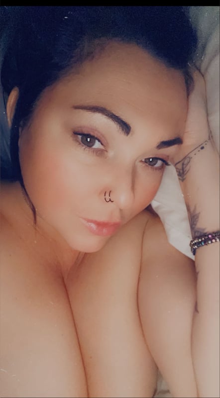 A woman with dark hair and tattoos poses lying down on a bed, smiling softly at the camera.