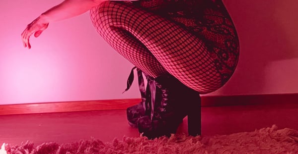 A person kneeling in patterned stockings and high boots against a pink-lit wall with a fluffy rug.