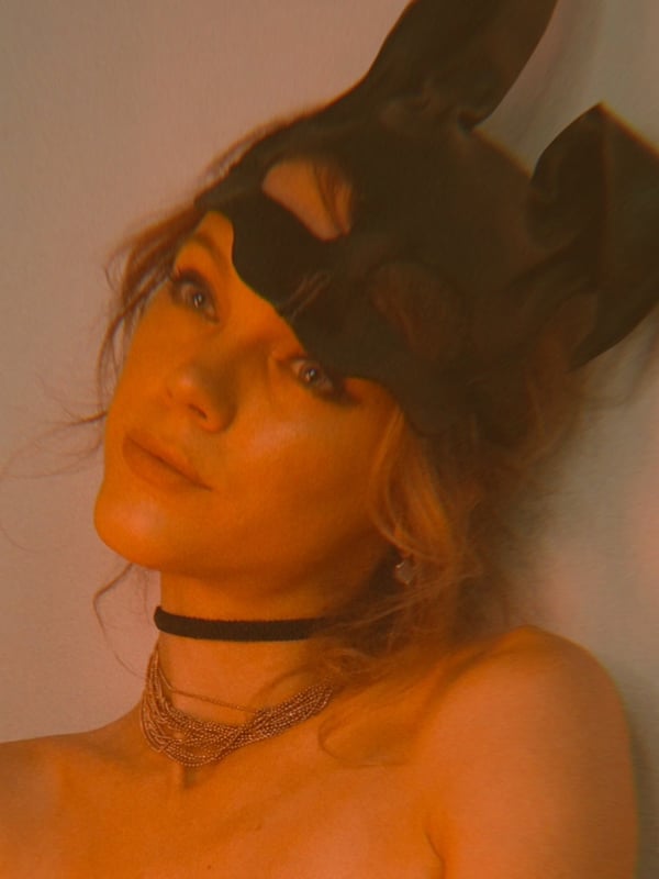 Close-up of a woman wearing a cat ears mask and choker necklace, posing with her head tilted against a wall.