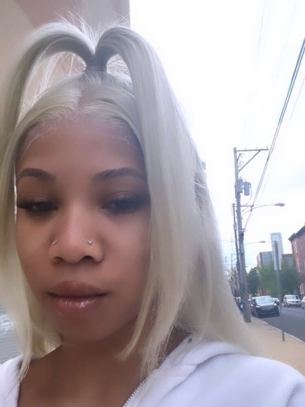 A young woman with platinum blonde pigtails, wearing a white hoodie, standing on a street with urban buildings and cars in the background.