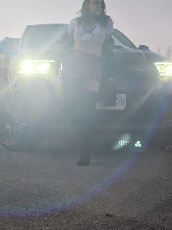 Woman leaning against a car with headlights on, posing confidently in a dimly lit outdoor setting.