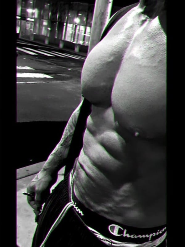 Muscular man standing shirtless on a dimly lit urban street at night, showcasing defined abs and arms.