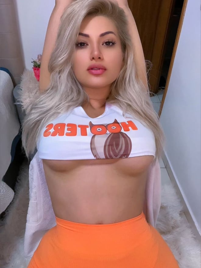 A woman with long blonde hair wearing a partially open branded top and orange bottoms, posing with arms raised in an indoor setting.