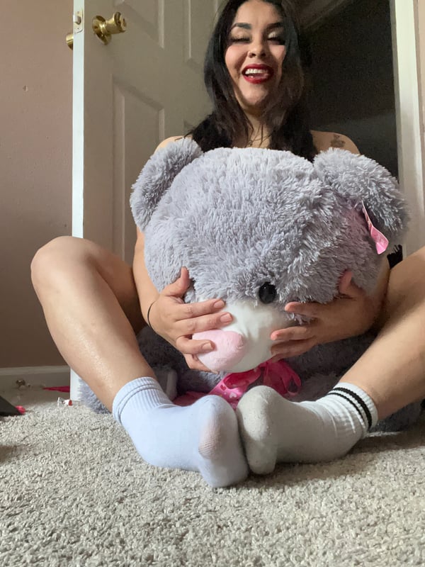 A smiling woman sitting on the floor holding a large gray teddy bear in a room with a door in the background.