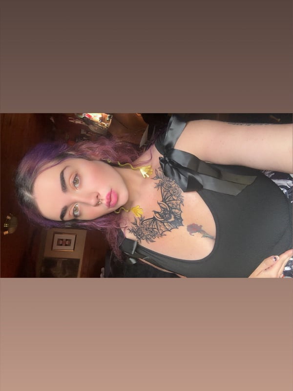 A young woman with purple hair and tattoos on her chest, wearing a black top, posing indoors with a neutral expression.