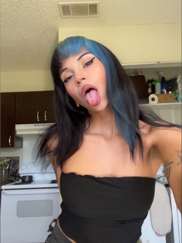 A young woman with black hair featuring blue streaks, sticking out her tongue while posing playfully in a kitchen, wearing a black tube top and pants.