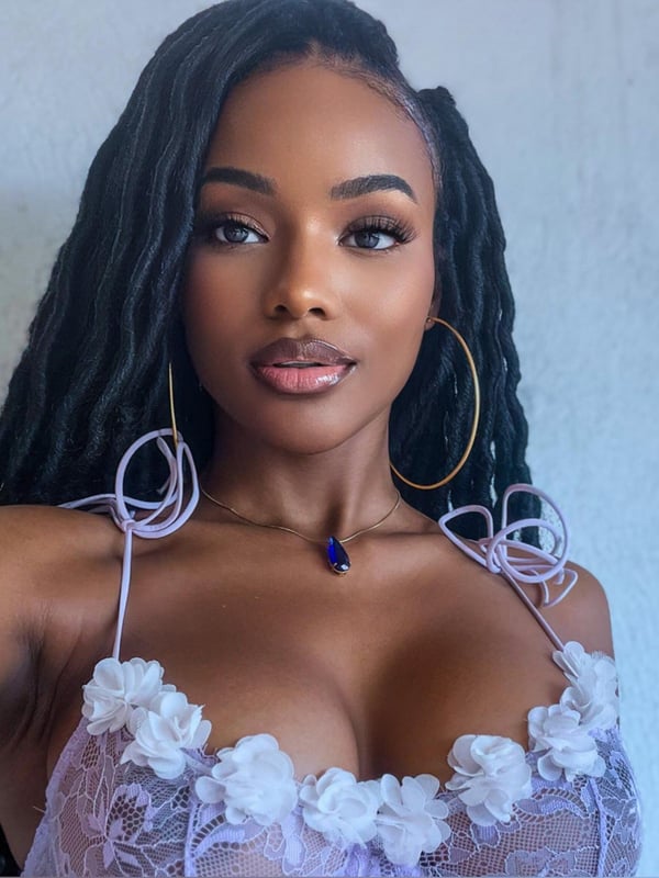 Close-up portrait of a woman with braided hair and hoop earrings, posing confidently in lace top against a plain background.