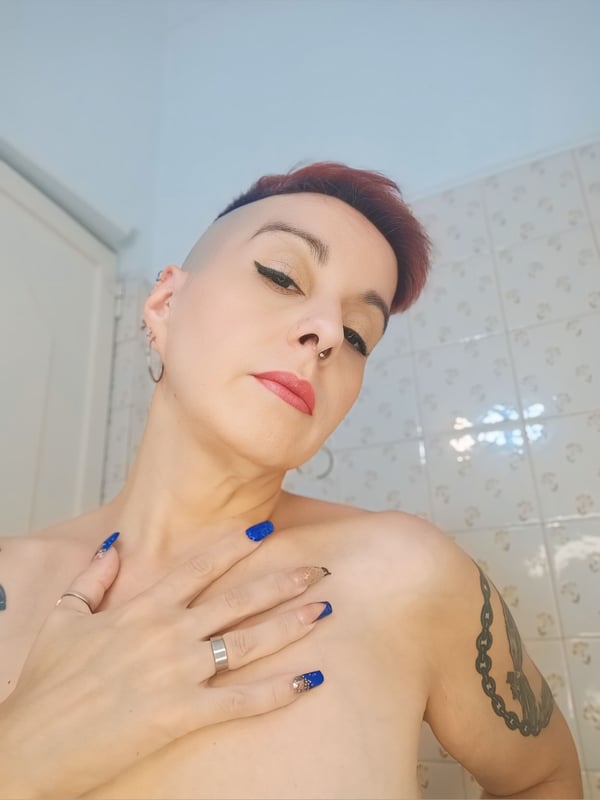 A woman with short red hair, blue nails, and tattoos poses topless in a bathroom, hand on her chest, wearing earrings.