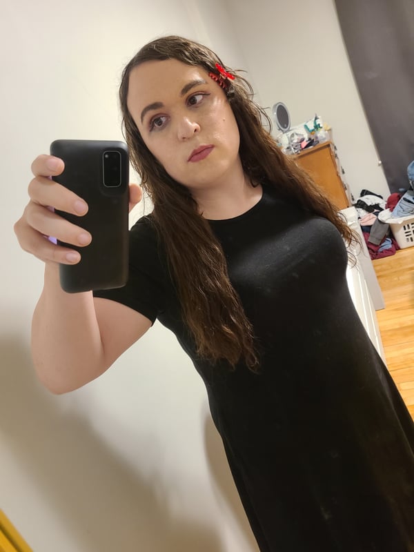 A person with long brown hair wearing a black dress, taking a mirror selfie indoors.