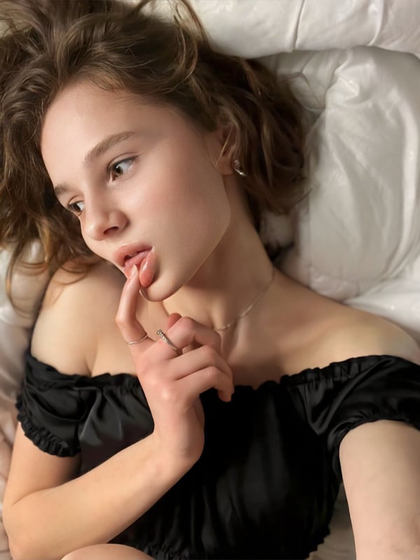 Young woman with wavy brown hair lying on a white bed in an off-shoulder black top, touching her lips thoughtfully.