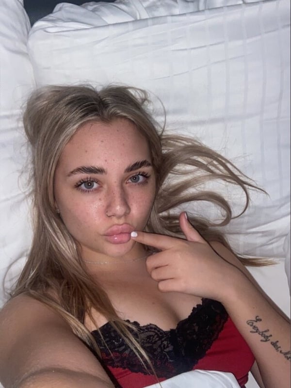 Young woman with long blonde hair lying on white sheets on a bed, wearing a black lace top, making a playful gesture with her finger to her lips.