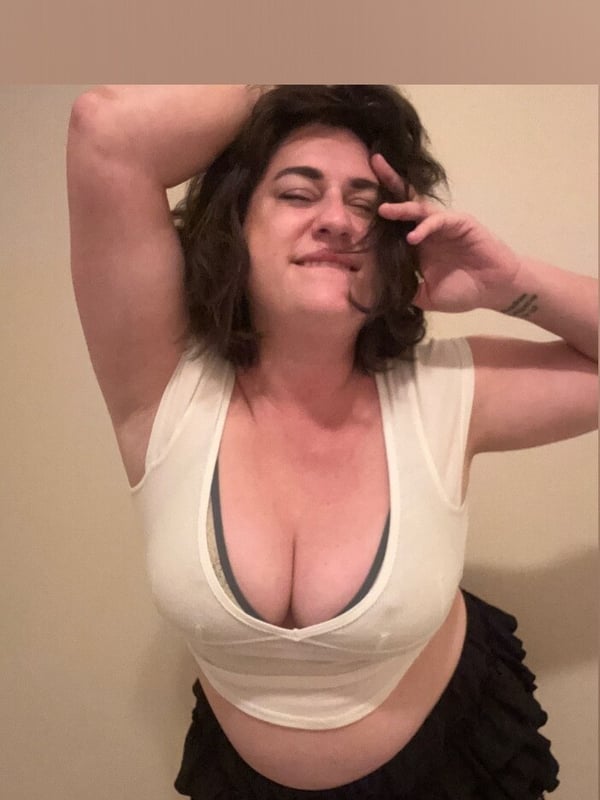 A woman with curly brown hair posing playfully with arms raised, wearing a white cropped top and black frilled skirt against a neutral background.