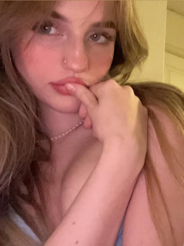 Young woman with long wavy hair sitting on a bed, touching her lips thoughtfully while wearing a necklace in a cozy indoor environment.