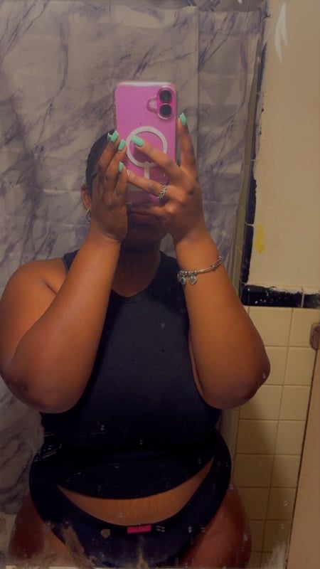 A woman taking a mirror selfie in a bathroom, covering her face with her hands while holding a pink phone, dressed in a black tank top and shorts.