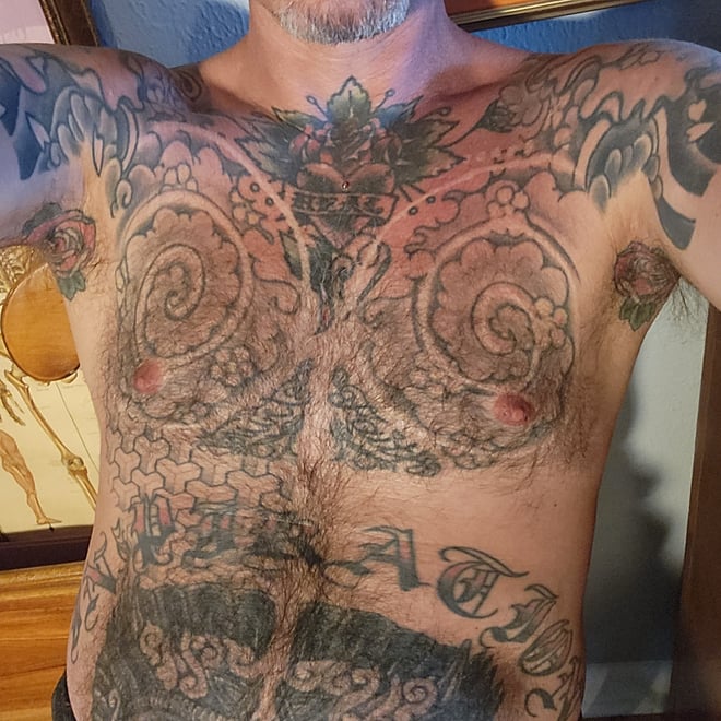 A shirtless man with extensive tattoos and body hair posing indoors near a window and artwork.