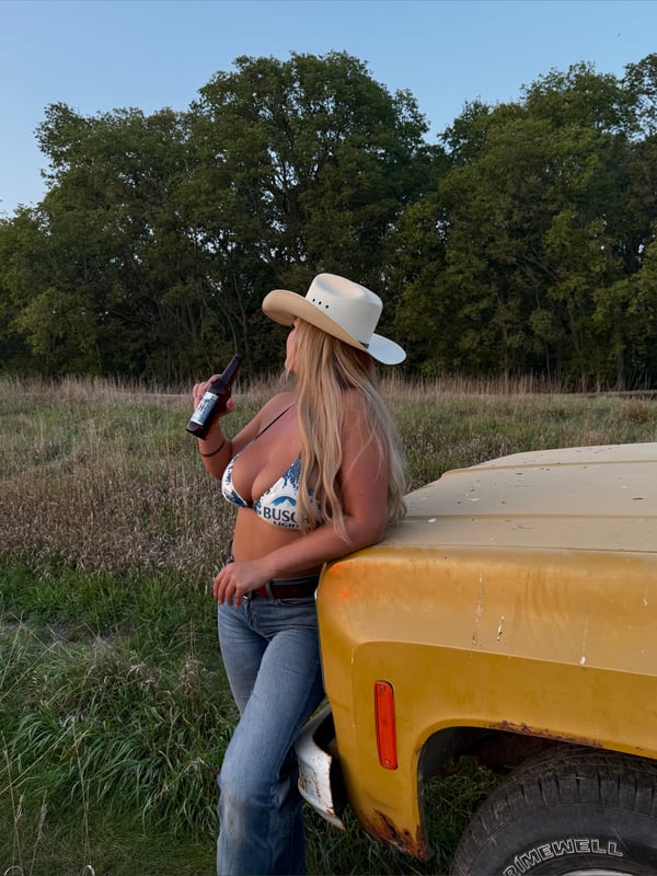 A woman in a cowboy hat and bikini top leaning against a yellow truck in a grassy field, holding a bottle.