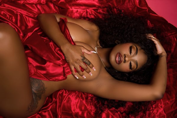 A woman with curly hair reclining on red silk sheets, posing elegantly with one hand raised.