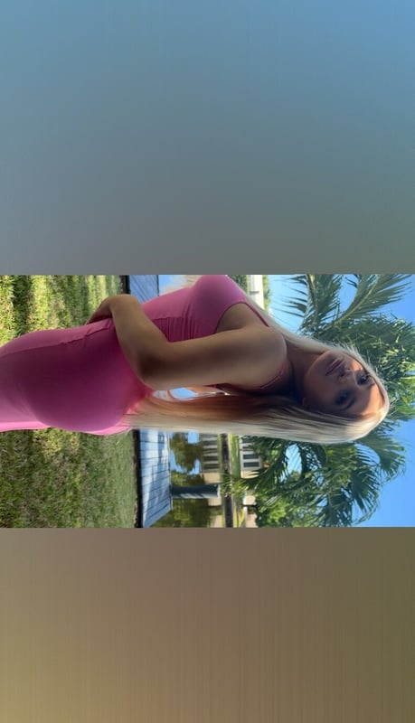 A person in a pink outfit posing sideways outdoors near palm trees and a pool with a scenic tropical background.