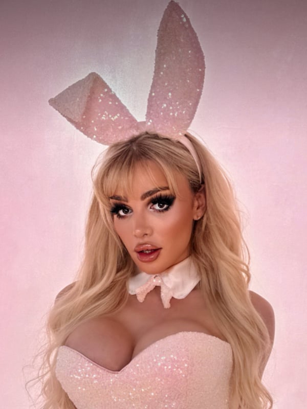 A person wearing a sparkly pink bunny outfit with ears and collar, posing playfully against a pink background.