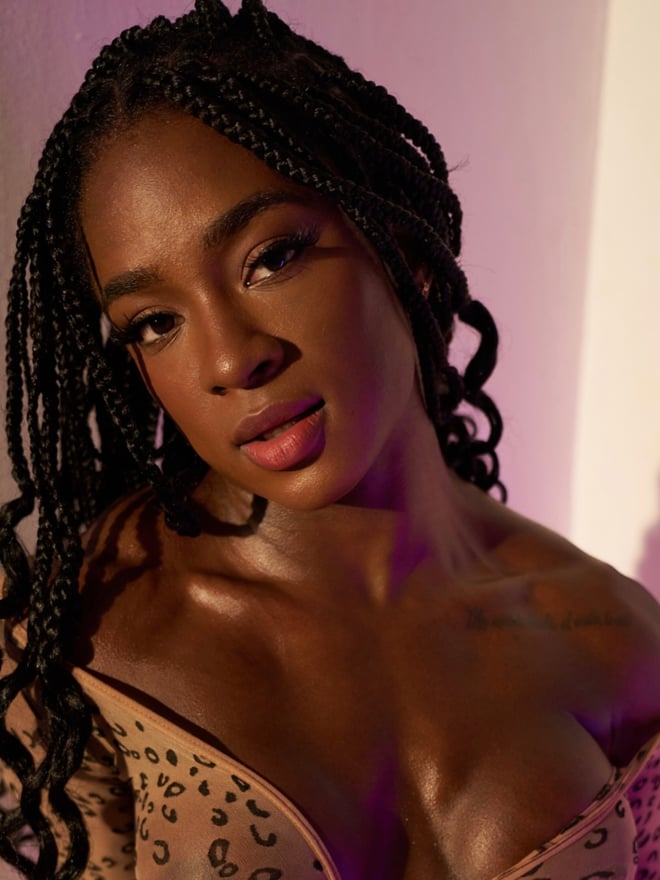 Close-up portrait of a woman with braided hair wearing a leopard print top, posing confidently against a purple background.