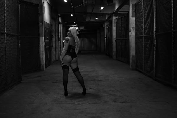 A woman with long blonde hair poses confidently in a black outfit and stockings in an industrial hallway with chain-link fencing and dim lighting.