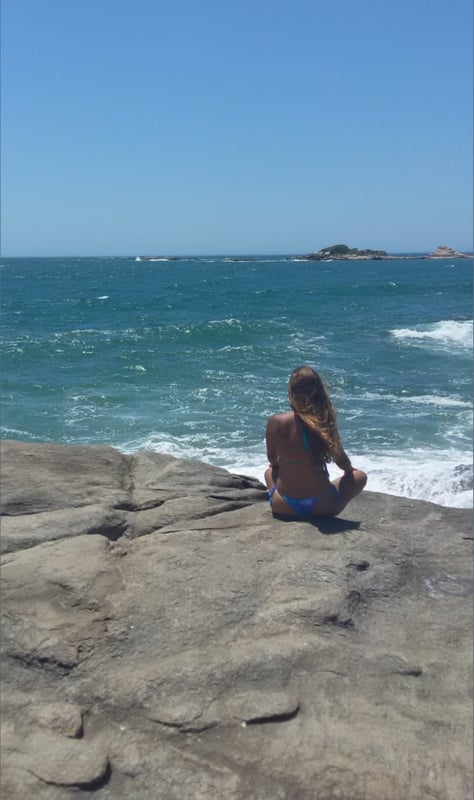 A woman with long hair sits on rocks by the ocean, facing away toward the blue sea and distant islands under a clear sky.