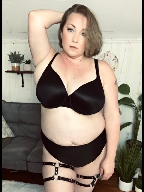 Woman standing confidently in a cozy indoor setting with plants, wearing a black top and bottoms with decorative straps.
