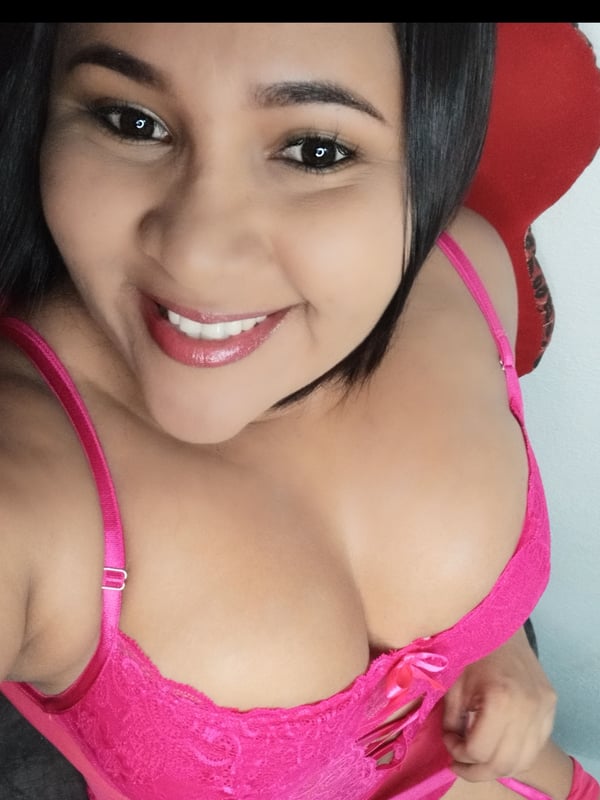 Close-up portrait of a smiling woman with dark hair, wearing a pink top, against a simple indoor background.