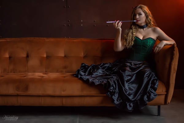 Woman with long wavy red hair wearing a green corset and black skirt, lounging on a brown velvet couch in a dimly lit room.