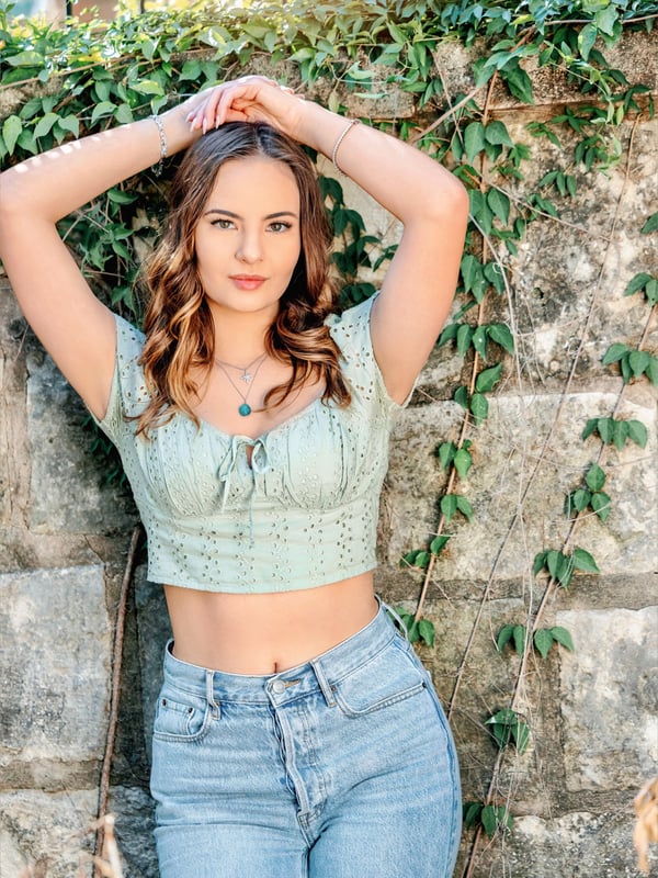 A young woman standing outdoors against a stone wall covered in green ivy, wearing a light green off-shoulder top and jeans.