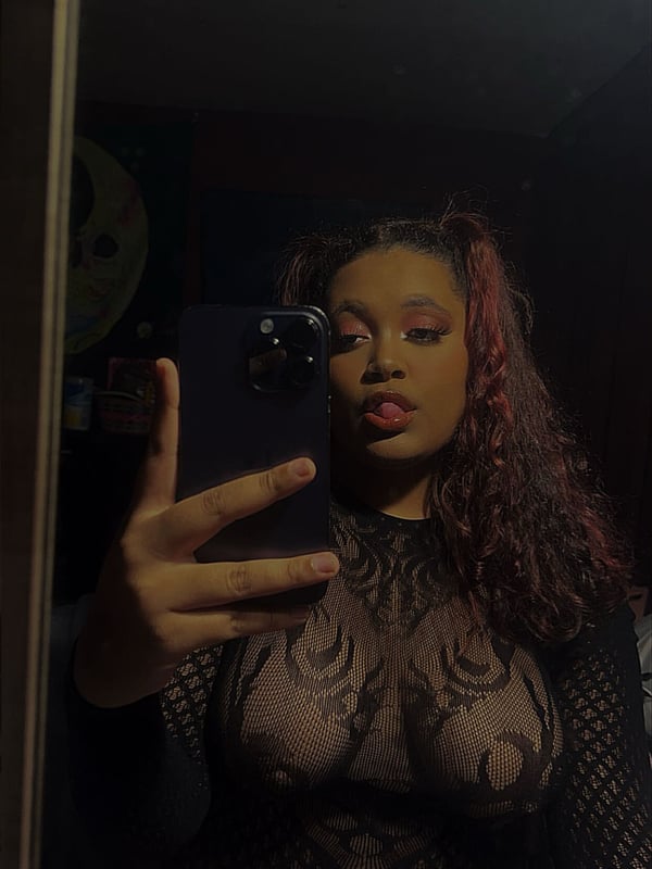 A woman taking a mirror selfie in a dimly lit room, wearing a black lace bodysuit and holding her phone.