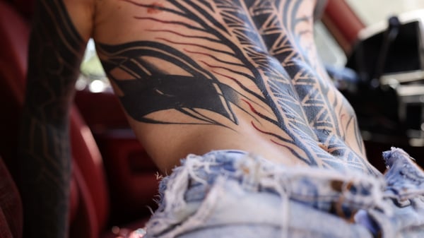 Close-up of a person's tattooed back and arms leaning against a car interior with worn jeans.