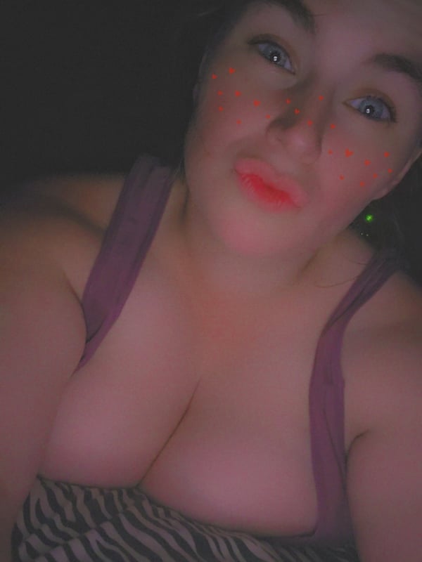 A woman with blue eyes and red freckles on her face, smiling while wearing a purple top in a low-light setting.