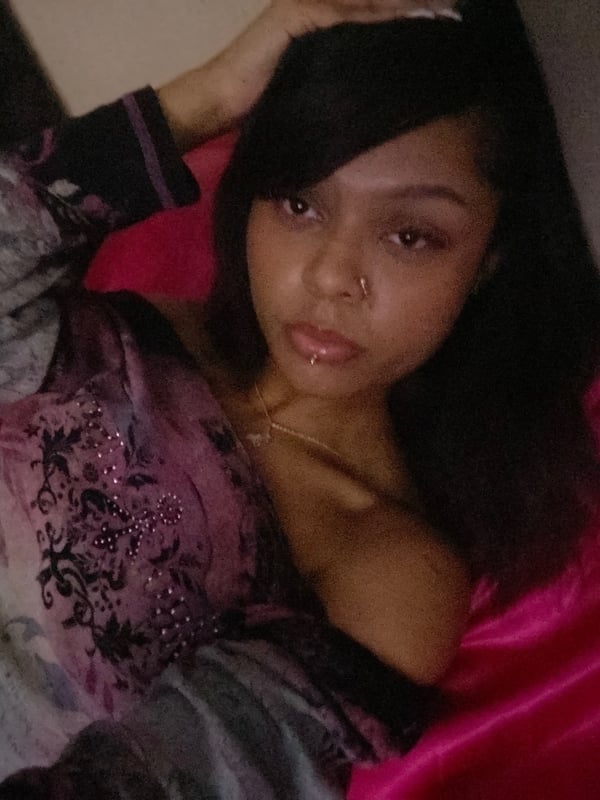 A young woman with long dark hair sitting on pink sheets, wearing a sparkly top, and smiling softly while leaning back.